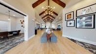 This impressive single-story amenity space is defined by its dramatic architectural centerpiece: a soaring vaulted ceiling with rich, dark-stained exposed wood beams running the full length of the main hall. A sculptural chandelier anchors the grand entry, casting warm light across light-toned hardwood floors that extend throughout the open-plan layout.

The expansive main area transitions seamlessly between distinct functional zones. A welcoming lounge features comfortable gray sofas arranged around a shiplap-accented wall with a mounted flat-screen television, complemented by modern accent chairs and layered area rugs. An adjoining reading or social nook displays a curated gallery wall of framed artwork and is furnished with a navy sofa and leather accent chair.

The dining area is a standout, featuring a long communal wood table surrounded by woven rope-back chairs with navy cushions, all beneath striking industrial pendant lights and a bold geometric-patterned accent wall. A private meeting or conference room offers an intimate setting with a marble-top round table and leather seating, finished with original artwork and a patterned carpet.

Floor-to-ceiling windows at the entrance flood the space with natural light, highlighting the airy, open ambiance. The overall design balances contemporary furnishings with warm architectural details, creating a sophisticated and welcoming environment throughout.