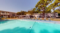 Located at 1002 Mayflower Dr in Wilmington, North Carolina, this outdoor amenity space centers around a generously sized swimming pool with clear, blue-green water. The pool is framed by a wide, open sun deck that provides ample room for lounging, with multiple reclining chairs arranged along all sides to accommodate residents seeking sun or shade. Safety lane lines are visible within the pool, and entry is provided via metal handrail steps. A chain-link fence encloses the perimeter, with a gated access point ensuring controlled entry to the area. One side of the deck features an additional shaded seating area with a table and umbrella, alongside a volleyball net, offering recreational variety beyond swimming. A utility or storage structure with a screened facade sits adjacent to the pool area, adding functional support to the space. Tall, mature pine trees border the area on multiple sides, providing a natural canopy that offers partial shade and a sense of privacy. The surrounding residential buildings, featuring balconies and exterior walkways, overlook the pool area. The space benefits from abundant natural light under open Carolina skies, creating a bright and inviting outdoor retreat well-suited for warm-weather enjoyment.