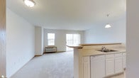 This single-floor space offers an open-concept layout that integrates the kitchen and living area into one cohesive, functional environment. The kitchen is outfitted with white cabinetry, a double-basin sink, a dishwasher, and light countertops that wrap around a peninsula with a raised breakfast bar. The peninsula serves as a natural divider between the kitchen and the expansive carpeted living area, maintaining an open and airy feel throughout.

The living space is finished with neutral light grey carpeting and crisp white walls, creating a clean, versatile backdrop suitable for a variety of furnishing styles. A wall-mounted HVAC unit provides in-unit climate control. Natural light enters generously through two windows on the far wall, alongside a large sliding glass door that brightens the entire space and provides access to the outdoors.

The overall ambiance is calm and understated, with a practical, no-frills design that maximizes usable space. The consistent neutral palette and open floor plan make this unit highly adaptable, offering a comfortable and functional living environment on a single level.
