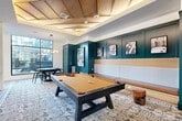 Located at 2500 Vantage Dr in Woodbridge, Virginia, this well-appointed game and lounge room delivers a sophisticated social atmosphere within a compact yet efficiently designed single-level layout. The space is anchored by a full-size pool table positioned at its center, surrounded by a richly patterned area rug that adds warmth and visual depth to the room.

One of the most distinctive architectural elements is the coffered wood ceiling, accented with ambient lighting that casts a warm glow throughout the space. Bold teal-painted paneled walls serve as a defining design feature, displaying a curated collection of black-and-white artwork that adds character and refinement. A built-in bench seat with a natural wood trim runs along the teal wall, offering relaxed seating for onlookers and guests.

A dramatic geometric-patterned feature wall in neutral gold and gray tones hosts dual mounted flat-screen televisions, creating a dedicated media zone. Adjacent to this, floor-to-ceiling black-framed glass walls and large windows invite abundant natural light and provide views to the surrounding exterior, visually expanding the room. Light hardwood flooring transitions into carpeted areas, and additional seating arrangements with bistro-style tables complete the lounge. The overall ambiance is modern, energetic, and thoughtfully designed for leisure and social engagement.