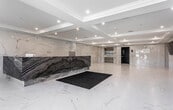Located at 1159–1183 Hand Pl in Elizabeth, NJ, this single-floor lobby space offers a refined and modern reception environment with high-end finishes throughout. The open layout provides a generous, unobstructed floor plan that immediately conveys a sense of spaciousness and professionalism upon entry.

The most commanding design element is the large reception desk, clad in bold, dramatically veined dark stone with sweeping grey and black wave-like patterns that contrast strikingly against the otherwise bright interior. Floor-to-ceiling wall cladding in white marble with subtle grey veining wraps the perimeter, lending the space a cohesive, polished aesthetic. The same white marble-style material extends seamlessly across the expansive floor, enhancing the sense of openness and cleanliness.

A coffered ceiling with recessed lighting illuminates the space evenly, adding architectural depth overhead. Multiple flat-panel displays are mounted on the marble walls, providing informational or visual content for visitors. Elevator access is visible toward the rear, alongside a secure entry door. Natural light filters in through windows near the entrance, complementing the bright, airy atmosphere. Entry mats at the front door provide a practical finishing touch to this elegantly designed lobby.