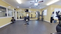 This fitness center offers a comprehensive single-floor layout designed to accommodate a full range of workout activities. The space is finished with warm yellow walls, crisp white crown molding, and durable rubber flooring throughout, creating a clean and energizing atmosphere.

The main workout area serves as the heart of the facility, featuring a wall-length mirror that visually expands the space and a wall-mounted flat-screen TV for entertainment during workouts. A ceiling fan with integrated lighting provides air circulation and overhead illumination, complemented by recessed lighting. This room is equipped with a multi-station cable machine, a dumbbell rack with a full range of free weights, a weight bench, and a barbell setup — offering a thorough strength training experience. Natural light filters in through windows, adding to the open feel.

An adjacent cardio room houses multiple treadmills and an elliptical trainer, all from True Fitness, arranged along the walls for ample workout space. An upright stationary bike is also available in a transitional area, providing further cardiovascular options.

A separate, private tanning room features bold yellow walls, a full tanning bed, a standalone fan, and a full-length mirror — offering a relaxing amenity alongside the fitness offerings. A bulletin board area near the entrance provides informational resources for users.