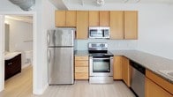 This two-floor space offers a clean, contemporary aesthetic characterized by light wood-style flooring, soft neutral wall tones, and exposed ceiling details that lend an industrial edge. The kitchen is well-appointed with warm honey-toned cabinetry, granite-style countertops, and a full suite of stainless steel appliances including a refrigerator, range, and built-in microwave. A peninsula counter provides a natural transition into the open living area, which benefits from wide windows that draw in natural light and offer views of the surrounding urban streetscape.

The layout includes multiple rooms across both floors, each finished with the same light flooring and crisp white trim that creates a cohesive, airy feel throughout. A wrought-iron staircase railing adds a refined architectural detail to the stairwell, connecting the two levels. Two full bathrooms serve the space — one features a combination tub and shower enclosed by sliding glass doors alongside a dark-vanity cabinet, while the second bathroom offers a similar functional layout. Exposed pipe runs and industrial ceiling elements contrast with the otherwise clean, modern finishes, adding character to the overall design.
