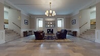This single-floor common space presents a refined, classical aesthetic that blends architectural tradition with functional design. The centerpiece of the space is a formal lounge area anchored by a decorative fireplace framed by a large mirror, flanked by symmetrically placed windows that allow natural light to fill the room. A brass chandelier with candle-style shades hangs above, casting warm light over a richly patterned area rug where leather armchairs and a plush sofa are thoughtfully arranged. Wainscoting lines the lower walls throughout, reinforcing the space's formal character, while crown molding and a soft neutral color palette lend a sense of understated elegance. Framed black-and-white photographs adorn the walls, adding a layer of historical character and local identity to the space. The entryway features a vaulted arched ceiling and a classic paneled door with wire-glass inserts, creating a welcoming and secure point of arrival. Adjacent to the main lounge, a dedicated mailroom offers a practical counterpart to the grandeur of the lounge, featuring a colorful mosaic tile floor in warm earth tones, built-in mailboxes, a sorting table, and additional framed artwork. Together, these spaces form a cohesive, well-appointed communal environment that balances classic architectural detail with everyday functionality.