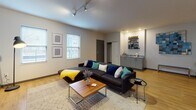 Located at 29 E Green St in Champaign, Illinois, this contemporary multi-floor residence offers a thoughtfully designed layout with modern finishes throughout. The open-concept kitchen is a standout feature, showcasing sleek gray cabinetry extending to the ceiling, stone countertops, a white subway tile backsplash, stainless steel appliances including a refrigerator, range, microwave, and dishwasher, and light hardwood-style flooring. A kitchen island provides additional prep and dining space, complemented by pendant lighting overhead. The living area is bright and spacious, featuring track lighting, multiple windows that invite natural light, and a built-in wet bar or kitchenette nook — ideal for entertaining. The bathrooms are finished with gray vanity cabinetry, stone-topped counters, large mirrors, and walk-in glass-enclosed showers with wood-look tile surround. Bedrooms are comfortably sized with carpeted floors, ample closet space, and windows offering outdoor views, including glimpses of a pool area. Additional utility space accommodates storage needs. The overall aesthetic is clean, modern, and functional, with a consistent palette of neutral tones, warm wood flooring, and contemporary fixtures carried throughout the residence.