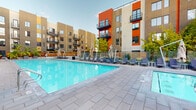 Misora's outdoor amenity area is a thoughtfully designed communal space centered around a generously sized, crystal-clear resort-style pool. The pool deck is finished with large-format stone pavers that provide a clean, contemporary aesthetic, complemented by an array of lounge chairs and patio seating arranged along the perimeter for relaxation and socializing. A dedicated spa area, with a capacity of four, is accessible via a glass-door entrance along the building's ground-floor facade, offering a more intimate retreat. Lush tropical plantings in oversized planters add vibrant greenery and a resort-like ambiance to the deck, softening the clean architectural lines. The space is enclosed by the surrounding multi-story building, which features a striking exterior palette of warm gold and bold orange panels, with private balconies overlooking the courtyard below. The building's modern design incorporates large windows and clean geometric forms, creating a dynamic urban backdrop. The Misora entry point to the amenity level is marked by a branded entry mat and seating near glass-paneled doors, offering a welcoming transition into the outdoor space. The overall atmosphere is vibrant yet relaxed, ideal for outdoor leisure in an urban residential setting.