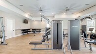 This fitness center offers a spacious, single-floor layout generously appointed with a full range of professional-grade exercise equipment. The interior design is clean and modern, featuring light hardwood-style flooring, crisp white walls with subtle decorative wainscoting, and tray ceilings accented with crown molding and recessed lighting. Ceiling fans are strategically positioned throughout to ensure comfortable air circulation during workouts.

The strength training area is anchored by multiple Life Fitness cable and selectorized machines, a power rack with weight plates, and a tiered dumbbell rack stocked with a wide range of free weights, accompanied by adjustable workout benches. The cardio section features treadmills and a recumbent bike, all positioned near large windows that allow abundant natural light to fill the space.

Wall-mounted flat-screen televisions are placed at multiple vantage points throughout the room, providing entertainment during workouts. Large mirrors along the walls enhance the sense of openness while serving a practical function for form monitoring. Stability balls and additional accessories are neatly stored along the perimeter. A decorative glass-panel entry door adds an architectural touch to the space, contributing to its polished and welcoming ambiance overall.