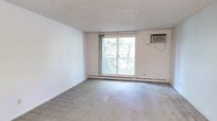 This single-floor residence offers a practical and comfortable layout with distinct living and kitchen areas. The living room is finished with wall-to-wall carpet and a textured ceiling, creating a quiet, insulated atmosphere. A sliding glass door fills the space with natural light and provides direct access to a private balcony, where black metal railings frame a pleasant view of mature trees and greenery below—an appealing outdoor retreat.

The kitchen is accessible through an open doorway from the living area and features rich, dark wood cabinetry with raised panel detailing spanning both upper and lower sections. Ample counter space with dark laminate countertops wraps in a U-shaped configuration, and a double stainless steel sink is positioned conveniently along one side. White appliances, including a refrigerator and dishwasher, complement the cabinetry, while the hardwood-style flooring adds a practical, easy-to-clean surface.

The living room is also equipped with a wall-mounted air conditioning unit and baseboard heating, ensuring year-round comfort. The overall design is straightforward and functional, with neutral wall tones throughout lending a clean, versatile canvas.