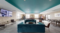 Located at 697 Bronx River Rd in Yonkers, NY, this dynamic amenity lounge is a single-floor recreational space designed to encourage socializing and entertainment. The room's most striking architectural feature is its bold teal ceiling, which adds a vivid pop of color against the neutral taupe walls and light grey carpeting, creating a lively yet cohesive aesthetic throughout.

The space is thoughtfully divided into distinct activity zones. A central lounge area anchors the room with a pair of tufted teal velvet sofas arranged around a coffee table, offering a comfortable gathering spot for residents. Nearby, a large flat-screen television is mounted on the wall, complemented by sleek accent chairs for additional seating.

The game zone is a standout feature, housing a regulation-style pool table with a red felt surface, classic upright arcade cabinets, and an immersive racing simulator — catering to a wide range of recreational interests. A high-top dining table with woven chairs provides a casual spot for dining or socializing.

Large windows wrap around multiple walls, flooding the space with natural light and offering clear views of the surrounding waterway, enhancing the open and airy atmosphere. A glass door provides direct outdoor access, further connecting the interior to its scenic waterfront setting. Recessed lighting throughout ensures the space remains well-lit at all times.