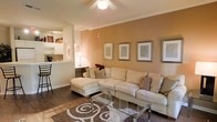 This single-floor residence offers a thoughtfully designed open layout that promotes a sense of flow and connectivity throughout the space. The living area serves as the heart of the home, anchored by a large sectional sofa in a neutral cream tone, complemented by a glass-top coffee table and a patterned area rug that adds texture and visual interest underfoot. Warm terracotta-toned accent walls create a cozy, welcoming ambiance, while crown molding along the ceiling adds a refined architectural touch. A ceiling fan provides both practical comfort and a clean focal point overhead. Framed artwork lines the accent wall, lending a curated, gallery-like aesthetic to the room. The living area opens directly to a kitchen and dining nook, visible through a pass-through opening that enhances the open-concept feel. The kitchen features light cabinetry and counter seating, making it equally functional for casual dining. Dark hardwood-style flooring runs throughout, grounding the warm color palette and tying each area together cohesively. Overall, the space balances comfort and style with a harmonious blend of warm tones, tasteful décor, and practical design elements suited for everyday living.