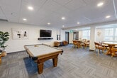Located at 301 Sandwich St N in Amherstburg, Ontario, this well-appointed amenity lounge offers a thoughtfully designed communal space across a single floor. The open-concept layout accommodates a variety of social and recreational activities, making it a versatile gathering hub.

The games area is anchored by a solid wood billiards table set atop patterned carpet tiles, complemented by a wall-mounted flat-screen television and a pair of cushioned armchairs — perfect for casual socializing. Adjacent to this is a dedicated dining nook featuring a round wood table surrounded by warm-toned chairs, ideal for small group gatherings.

A comfortable lounge area offers generous sectional seating, a wall-mounted television, and tasteful décor including framed artwork and accent pieces, creating a relaxed, living-room atmosphere. Natural light filters in through vertical-blind windows, enhancing the bright, neutral interior.

The kitchenette is a standout feature, showcasing rich dark wood cabinetry, a light granite-topped breakfast bar with upholstered bar seating, stainless steel appliances including a full-size refrigerator, and a coffee station — all finished with a clean tile backsplash. Recessed lighting throughout the space maintains a warm and inviting ambiance, while the consistent neutral palette and quality finishes lend the entire lounge a polished, modern aesthetic.