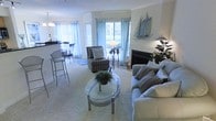 This single-floor residence offers an open, flowing layout that connects the living area to a dining and kitchen space. The living room serves as the focal point, featuring a fireplace with a clean white surround that anchors the room. Soft, neutral-toned carpeting extends throughout, complementing the light, airy color palette of the space. A glass-top oval coffee table with a metallic frame sits at the center of the seating arrangement, which includes a plush sofa adorned with striped accent pillows and a coordinating armchair. A standout decorative element is the large sculptural sailboat mounted on the wall, lending a nautical character to the space. Multiple windows dressed with sheer teal curtains and white blinds allow generous natural light to fill the room. Adjacent to the living area, a raised countertop bar connects to the kitchen and dining area, where bar-height seating and a chandelier are visible. The overall ambiance is calm and coastal, with carefully chosen decor and a cohesive soft color scheme creating a welcoming, comfortable atmosphere throughout.