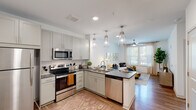 This single-story residence showcases a thoughtfully designed open-concept layout that seamlessly connects the kitchen, dining, and living areas. The kitchen is a standout feature, equipped with stainless steel appliances including a range, microwave, refrigerator, and dishwasher. Soft gray shaker-style cabinetry pairs elegantly with dark granite countertops, while a peninsula island provides additional prep space and casual seating. Three pendant lights with geometric glass shades hang above the island, adding a modern touch. Wood-look flooring flows throughout the main living areas, grounding the bright, white-walled interior.

The living room benefits from generous natural light through large windows dressed with sheer curtains, and a ceiling fan ensures year-round comfort. The bedroom is a tranquil retreat featuring plush carpet, a ceiling fan, and windows that invite natural light. An en-suite bathroom includes a granite-topped vanity with undermount sink, gray cabinetry, a glass-enclosed walk-in shower with tile surround, and a large mirror with contemporary lighting. A dedicated entryway with recessed lighting provides a clean transition into the main living space. Overall, the residence presents a cohesive, contemporary aesthetic throughout.