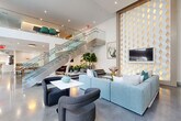 This elegant two-story amenity space is designed with a modern aesthetic that balances comfort and sophistication throughout. The ground floor welcomes visitors with a soaring double-height lobby area bathed in natural light from expansive floor-to-ceiling glass walls that frame views of an outdoor pool courtyard. Polished concrete floors run throughout, anchoring the contemporary design.

The main lounge area features plush light-blue sectional sofas, sculptural armchairs, and curated artwork, all set beneath soaring ceilings adorned with a striking gold diamond-patterned decorative wall panel. A casual dining and banquette area showcases a long teal upholstered bench seat lined with bold portrait artwork and paired with bentwood chairs and bistro tables.

A fully equipped catering kitchen provides stainless steel appliances, including a side-by-side refrigerator, built-in ovens, and a microwave, all complemented by light wood cabinetry and a distinctive sage-green elongated tile backsplash. A generous white waterfall island with pendant lighting and leather barstools completes the kitchen.

A glass-railed staircase leads to the second floor, where a mezzanine overlooks the main level and offers additional lounge and meeting space. Large windows on both floors ensure the entire space remains bright and connected to its surroundings.