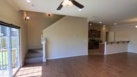 This two-story home offers a well-designed open-concept layout that seamlessly connects the main living spaces. The living area is anchored by rich, dark hardwood flooring and features vaulted ceilings that lend an airy, expansive feel to the space. A ceiling fan with a light fixture is centrally positioned, complementing the recessed lighting that illuminates the room. Neutral-toned walls provide a versatile backdrop throughout. A carpeted staircase with clean white balusters leads to the upper floor, adding a classic architectural detail to the open layout. The kitchen is visible from the living area and is outfitted with warm wood cabinetry, stainless steel appliances including a range and microwave, and a breakfast bar that serves as both a functional workspace and casual dining area. The transition between the living room and kitchen is defined by decorative columns, creating a sense of separation while maintaining the open flow. Natural light filters in through windows along the lower level, brightening the main floor. The overall ambiance is clean, modern, and move-in ready, with a cohesive design that balances comfort and practicality across both levels of the home.