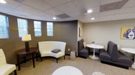 This single-floor commercial space is thoughtfully designed to offer a comfortable and professional environment. The lounge area serves as the focal point, featuring a curated mix of seating arrangements that balance style and functionality. A tufted white curved settee adds an elegant touch, complemented by charcoal gray upholstered seating and a sleek round pedestal table — ideal for casual meetings or relaxed conversation.

The interior is finished with warm taupe and gray walls that create a calm, neutral backdrop, while patterned carpeting adds texture and visual interest underfoot. A drop ceiling with recessed lighting provides even, ambient illumination throughout the space. Narrow horizontal windows along one wall allow natural light to filter in, enhancing the overall brightness without compromising privacy.

Decorative accents, including wall art and a small potted plant, contribute to the polished yet approachable atmosphere. The thoughtful layout encourages both individual comfort and small group interaction, making this space well-suited for use as a waiting area, client lounge, or collaborative meeting space. Overall, the design strikes a balance between modern professionalism and inviting warmth.