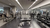 This expansive fitness center is thoughtfully designed to accommodate a full range of workout routines across clearly defined training zones. The facility is laid out on a single floor, with a generous open floor plan that provides ample space between equipment stations, promoting a comfortable and unobstructed workout experience.

The cardio zone stretches along a wall of large floor-to-ceiling windows, allowing abundant natural light to fill the space while offering views of the surrounding greenery. This area is lined with a row of treadmills, elliptical trainers, stationary bikes, and stair climbers from reputable brands including Precor, each equipped with individual display monitors.

Transitioning deeper into the facility, the strength training zone features an extensive selection of cable machines, multi-functional selectorized weight stations, and free weight areas stocked with dumbbells, barbells, and adjustable benches. Full-length mirrors are strategically mounted along the walls to assist with form and create a sense of added depth. Exposed concrete structural columns add an industrial character that contrasts with the clean white walls and modern drop-tile ceiling fitted with recessed and panel lighting.

The flooring throughout consists of durable speckled rubber and vinyl composite, providing both safety and resilience. A functional training area accommodates suspension trainers, resistance bands, and additional specialty equipment. The overall ambiance is clean, modern, and professional.