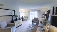 This single-floor residence features an open-concept layout that seamlessly connects the living and dining areas, creating a spacious and functional environment. The living area is finished with plush carpeting and centers around a glass-top coffee table, with comfortable seating arranged to encourage conversation. A decorative credenza with textured paneling and ring hardware anchors one wall, adding a stylish focal point alongside a framed mirror that enhances the sense of depth and light.

The dining area is defined by a dark wood table paired with upholstered chairs in a contrasting light green tone, offering a refined yet casual space for meals. Large sliding glass doors dominate the far wall, flooding the interior with natural light and providing direct access to an outdoor balcony or patio. Sheer white curtains frame the doors, softening the transition between indoors and outdoors while preserving the airy atmosphere.

The neutral color palette throughout—whites, grays, and warm taupes—creates a cohesive and calming ambiance. Overall, the space feels well-proportioned, bright, and thoughtfully arranged for everyday living.