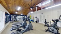 This fitness center occupies a generously sized single-floor space designed to accommodate a full range of workout activities. The most striking architectural feature is the vaulted, tongue-and-groove wood-plank ceiling, which lends a warm, lodge-like character to the room while significantly enhancing the sense of vertical space. A large ceiling fan is centrally mounted, promoting air circulation throughout the facility. Natural light enters through clerestory windows positioned high along the walls, supplemented by suspended fluorescent fixtures.

The interior is finished with neutral carpet flooring and crisp white walls accented by bold navy blue sections, creating a visually engaging and energetic atmosphere. Strategically placed mirrors along multiple walls expand the perceived size of the space and allow users to monitor their form during exercise.

Cardio equipment is well-represented, with multiple treadmills, elliptical trainers, and stationary bikes arranged in organized rows. A dedicated functional training area features a rack of stability balls in various sizes, medicine balls, resistance bands, and balance training tools such as a BOSU ball and foam rollers. A multi-use weight bench and dumbbell rack round out the strength training options. French doors provide secondary access and enhance natural light. Overall, the space maintains a clean, functional, and inviting environment.