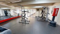 Located at 1111 Cardenas Dr SE in Albuquerque, New Mexico, this fitness center offers a well-organized and fully equipped workout space on a single floor. The room is designed to maximize functionality, featuring durable rubber flooring throughout that provides both safety and comfort during exercise. Large sliding glass windows and a glass door span much of one wall, flooding the space with natural light and offering views of the surrounding outdoor stairway and courtyard. A full-length wall mirror with a bold red accent stripe along one side enhances the sense of openness and allows users to monitor their form during workouts. The equipment layout is thoughtfully arranged to accommodate a variety of fitness activities. Cardio machines include a treadmill, elliptical trainer, stationary upright bike, and a recumbent bike. Strength training is supported by a multi-function cable machine, a power tower for bodyweight exercises, a Hoist adjustable bench, a dumbbell rack, and a freestanding punching bag. Informational fitness posters are displayed on the walls for reference. Overhead fluorescent lighting supplements the natural light, ensuring the space remains bright throughout the day. The textured ceiling and exposed utility lines give the room a practical, no-frills character well-suited to its purpose as a community fitness facility.