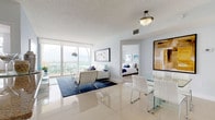 Suite 2412 is a thoughtfully designed high-rise residence spanning a single floor, offering an open and airy layout bathed in natural light. The living and dining area serves as the heart of the suite, featuring large floor-to-ceiling windows and sliding glass doors that frame stunning panoramic water and city views. Gleaming large-format tile flooring flows throughout the main living spaces, enhancing the sense of openness. A glass dining table with modern white chairs sits adjacent to a fully equipped kitchen, which features white cabinetry, granite countertops, stainless steel appliances, and a breakfast bar with contemporary bar seating — ideal for casual dining. The primary bedroom is a serene retreat, showcasing expansive windows with breathtaking elevated views, plush carpeting, and direct access to a well-appointed en-suite bathroom complete with a glass-enclosed walk-in shower and neutral tile finishes. The second bedroom offers carpeted flooring, a tufted headboard, and city-facing windows that bring in ample natural light. Soft, neutral tones throughout the suite create a cohesive and calming ambiance, making Suite 2412 a refined and comfortable high-floor residence.