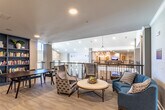 This thoughtfully designed communal space combines a curated library, comfortable lounge areas, and corridor access to additional rooms, all unified by a clean, modern aesthetic. The library corner is anchored by a dramatic floor-to-ceiling built-in bookshelf finished in a rich navy blue, filled with an extensive collection of books and decorative accents. A dark wood writing desk and plush wingback chairs complete this scholarly nook, creating an inviting space for reading or quiet reflection.

Adjacent to the library, the open lounge area features an elegant seating arrangement including a curved navy sofa, patterned accent chairs, and a tufted round ottoman serving as a central coffee table. Light wood-look flooring runs throughout, complementing the neutral gray and white wall palette. Recessed lighting and a statement pendant chandelier provide warm, layered illumination throughout the space.

An open railing system separates the lounge from an open atrium corridor below, adding a sense of vertical depth and connectivity. Crown molding details lend a refined, upscale character to the hallways. Double glass French doors lead to additional multipurpose rooms beyond, hinting at further amenities. Artwork displayed along the corridors adds warmth and personality, making this a truly inviting communal environment.