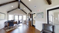Located at 133 Boone Ridge Dr in Johnson City, Tennessee, this single-story community or leasing office is designed to impress with its striking architectural details and welcoming atmosphere. The centerpiece of the space is its dramatic vaulted ceiling, accented by bold, dark-stained decorative wood beams that draw the eye upward and lend a warm, rustic-modern character to the interior. Dual ceiling fans keep the space comfortable throughout, while recessed lighting ensures bright, even illumination. Rich hardwood flooring runs throughout, complementing the crisp white wainscoting that lines the lower walls, creating a clean and polished aesthetic. The main open area serves as both a reception and meeting space, featuring a front desk workstation with dual monitors, comfortable seating for visitors, and a dedicated conference or meeting table near the rear. Large windows and glass-paneled double doors flood the space with natural light and offer views to an outdoor patio area. A compact kitchenette with dark wood cabinetry, a microwave, refrigerator, and coffee station provides added convenience. A community site map is prominently displayed on the wall, offering an informative overview of the surrounding property. The overall ambiance is professional yet inviting, blending contemporary design with warm, traditional touches.