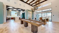 Located in Maricopa, Arizona, this single-floor community living room is a thoughtfully designed social hub that blends modern aesthetics with comfortable, functional spaces. The open-plan layout is anchored by soaring ceilings adorned with dark-stained exposed wood beam detailing, which creates a striking architectural contrast against the crisp white walls. Large-format travertine-style tile flooring flows seamlessly throughout, lending a cohesive, upscale feel to the entire space.

A standout shuffleboard table serves as a central focal point, positioned beneath an eye-catching arrangement of spherical woven pendant lights that add texture and warmth to the room. Floor-to-ceiling windows and glass doors flood the interior with natural light and offer clear views of the surrounding Maricopa community, creating a bright and inviting ambiance throughout the day.

Distinct seating zones cater to a variety of social settings. A generously sized built-in banquette with channel-tufted upholstery and accent pillows lines one sun-drenched window wall, while curated groupings of chairs and sofas provide additional conversation areas. A casual dining nook with a white countertop island, upholstered bar stools, and a cozy booth-style banquette — complete with decorative wall art and pendant lighting — adds a café-like atmosphere. The overall design is warm, modern, and highly social.