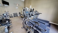 This fitness center offers a comprehensive workout environment within a single-level layout. The room is characterized by its vaulted ceilings, which lend an open, airy feel to the space, and neutral-toned walls that create a clean, focused atmosphere. Carpeted flooring runs throughout, providing comfort underfoot during workouts.

The cardio section is prominently featured and includes multiple treadmills, elliptical trainers, and a stair climber, each equipped with individual display consoles for tracking workout metrics. A multi-station cable and weight machine occupies a central area of the room, offering a variety of strength-training options. A dedicated free weights section lines one wall, featuring a well-organized dumbbell rack stocked with a range of weights to accommodate varying fitness levels.

A wall-mounted clock keeps users on schedule, while a pendant light fixture supplements the ambient lighting. Mirrors are positioned strategically toward the back of the room, enhancing the sense of depth and allowing users to monitor their form. Informational signage is posted on the wall, likely outlining facility rules and guidelines for equipment use.