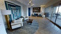 This single-floor space makes a strong visual impression through its carefully considered design elements and high-end finishes. The living area centers around a large circular area rug in soft grey tones, anchoring a grouping of white leather seating with gold-toned frames and a standout live-edge wood coffee table with resin detailing—an artisan centerpiece that draws the eye immediately.

A dramatic large-scale geometric artwork in deep blues and teals dominates one wall, complemented by a tall marble accent panel featuring bold veining in grey and gold tones. The blue color story continues with a sleek, contemporary sofa dressed in coordinating blue and teal cushions, positioned against a built-in wooden shelving unit displaying an artfully curated collection of decorative vessels and objects.

Light hardwood flooring in a herringbone-inspired layout adds warmth and texture to the space, while a sculptural multi-arm chandelier with gold-toned hardware provides a refined focal point overhead. Recessed lighting supplements the overall illumination. A mirrored panel along one wall subtly expands the sense of space. The overall ambiance is polished, design-forward, and cohesive.