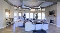 This beautifully appointed single-floor space makes an immediate impression with its distinctive coffered ceiling, where geometric wooden beams intersect in an octagonal pattern, adding architectural depth and visual interest overhead. A tasteful drum pendant chandelier anchors the center of the room, complementing the recessed lighting that illuminates the space evenly throughout.

Floor-to-ceiling windows wrap around the lounge area, flooding the interior with abundant natural light and creating an open, airy atmosphere. The expansive arrangement of white slipcovered sofas and seating clusters encourages conversation and relaxation, accented by navy and patterned throw pillows that introduce a refined coastal aesthetic.

Tasteful decorative touches — including tall potted fiddle-leaf fig trees, ornate candelabras, and curated tabletop arrangements — lend the space a polished, welcoming character. Dark console tables provide elegant contrast against the lighter furnishings, while a patterned area rug grounds the central seating zone.

The flooring features large-format stone-look tile in warm neutral tones, adding a sense of solidity and sophistication underfoot. Together, the architectural details, abundant glazing, and carefully curated furnishings create a bright, inviting atmosphere that is both functional and refined.