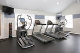 This fitness center is designed to support a comprehensive workout experience within a single-floor layout. The space is distinguished by vaulted ceilings that lend an open, airy feel, complemented by a two-tone paint scheme of soft blue and white that gives the room a clean, modern aesthetic. Dark rubber flooring runs throughout, providing a durable and practical surface suited for all types of exercise.

The cardio area is outfitted with multiple Matrix treadmills and an elliptical trainer, all positioned in front of large windows with slatted blinds that allow ample natural light to filter in while maintaining privacy. A wall-mounted flat-screen television is positioned for easy viewing during workouts.

The strength training zone features a multi-station cable machine, a dedicated weight rack, and additional resistance equipment, offering a variety of options for full-body conditioning. A body weight scale and informational signage are also present near the entry area, adding a functional touch.

Access to the facility is provided through both a main entry door with glass panels and a secondary exit, ensuring convenient flow. Recessed and pendant lighting supplements the natural light, keeping the space well-illuminated at all hours. Overall, this fitness center provides a practical, well-appointed environment for a wide range of exercise routines.
