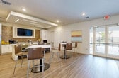 This 900 square foot space in Florida City showcases a modern, open-concept design that maximizes functionality and style. The interior is characterized by bright, airy spaces with a cohesive color scheme of white, yellow, and wood tones. The main area features a welcoming reception with the "Ambar Key" signage, setting a professional tone. 

The space includes a well-equipped kitchenette with white cabinetry, stainless steel appliances, and a brick-textured accent wall. A comfortable lounge area is furnished with plush seating in neutral tones, accented by vibrant yellow pillows. The design incorporates both practical and aesthetic elements, such as a large circular mirror and contemporary artwork.

A notable feature is the floor-to-ceiling glass doors that flood the space with natural light and offer views of an outdoor area with a pool. The flooring throughout is a warm, wood-like material that adds to the inviting atmosphere. Recessed lighting and sleek fixtures contribute to the modern ambiance.

The layout accommodates various functions, including a fitness area visible through interior windows, suggesting a multi-purpose use of the space. Overall, the design emphasizes a clean, contemporary aesthetic with attention to detail in both form and function.