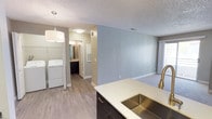 This single-floor unit presents a thoughtfully updated interior with a cohesive neutral color palette throughout. The kitchen serves as a functional centerpiece, featuring dark espresso-toned cabinetry, crisp white countertops, and a full suite of stainless steel appliances including a refrigerator, electric range, and built-in microwave. A peninsula with an undermount stainless sink and a brushed gold faucet separates the kitchen from the adjoining living area, providing a natural flow between spaces. The living area is carpeted and receives natural light through a sliding glass door that opens to a balcony, offering views of the surrounding Southwest-style buildings with terracotta tile rooflines and mature trees. Plank-style flooring runs through the kitchen and hallway areas, creating a clean transition between zones. An in-unit washer is conveniently located along the hallway. The bathroom includes a full tub-shower combination, textured walls, and a wall-mounted ventilation unit. Additional carpeted rooms provide flexible spaces, each finished with neutral walls, textured ceilings, and clean white baseboards throughout.