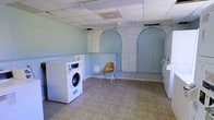 This single-floor utility space offers a functional laundry room with a distinctive character that sets it apart from typical utility areas. The room features light blue painted walls accented by decorative arched stone detailing, creating a surprisingly unique visual element within a practical setting. Two prominent arched recesses framed in stone masonry are built into the wall, adding architectural interest and depth to the space. A front-load washing machine is positioned along one wall, ready for use, while the open floor plan allows for comfortable movement and potential addition of further laundry appliances. The floor is covered in neutral tile, which is easy to maintain and well-suited to the demands of a laundry environment. A suspended drop ceiling with panel tiles runs overhead, providing a clean and even finish. The room offers ample open floor space beyond the appliances, making it practical for sorting, folding, or storing laundry supplies. The combination of the stone arch details and cool blue tones gives this utilitarian space a memorable aesthetic that blends character with everyday functionality.