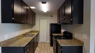 The kitchen is designed with a galley-style layout, maximizing efficiency through parallel runs of cabinetry and countertops along both walls. Dark espresso-toned upper and lower cabinets with brushed nickel hardware create a striking contrast against the light-toned walls and warm granite-style countertops, which provide ample prep space on both sides of the kitchen. The left side features a double-basin stainless steel sink with a standard faucet, accompanied by a dishwasher and additional lower cabinet storage. Overhead, upper cabinets extend toward the ceiling, offering generous storage capacity. On the right side, a black freestanding range with coil burners is paired with an overhead range hood, while a full-size black refrigerator anchors the end of the run. Flush-mount fluorescent lighting runs along the ceiling, ensuring the space is well-lit throughout. Light wood-tone flooring ties the kitchen together, adding warmth to the overall aesthetic. At the far end of the kitchen, a set of bifold doors likely conceals a pantry or utility closet, adding practical storage to this compact yet complete kitchen.