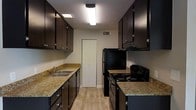 The kitchen is designed with a galley-style layout, maximizing efficiency through parallel runs of cabinetry and countertops along both walls. Dark espresso-toned upper and lower cabinets with brushed nickel hardware create a striking contrast against the light-toned walls and warm granite-style countertops, which provide ample prep space on both sides of the kitchen. The left side features a double-basin stainless steel sink with a standard faucet, accompanied by a dishwasher and additional lower cabinet storage. Overhead, upper cabinets extend toward the ceiling, offering generous storage capacity. On the right side, a black freestanding range with coil burners is paired with an overhead range hood, while a full-size black refrigerator anchors the end of the run. Flush-mount fluorescent lighting runs along the ceiling, ensuring the space is well-lit throughout. Light wood-tone flooring ties the kitchen together, adding warmth to the overall aesthetic. At the far end of the kitchen, a set of bifold doors likely conceals a pantry or utility closet, adding practical storage to this compact yet complete kitchen.