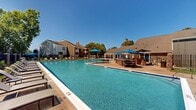 Located at 10303 7th Regiment Drive in Manassas, Virginia, this outdoor amenity area serves as a central gathering space for a multi-building residential apartment community. The centerpiece is a large, rectangular swimming pool with clear blue water, surrounded by an expansive pool deck designed for relaxation and recreation. The deck is lined with rows of adjustable lounge chairs, providing ample space for sunbathing along the pool's edges. Colorful blue patio umbrellas and additional seating areas are scattered throughout the deck, offering shaded spots for residents to gather. A charming pergola structure with a pitched roof sits adjacent to the pool area, providing a covered outdoor dining and seating space. The pool area is enclosed by a classic black wrought iron fence with brick pillar accents, which also borders a large, well-maintained open lawn area with lush green grass — ideal for outdoor activities. Mature trees provide natural shade throughout the grounds. The surrounding multi-story residential buildings feature brick and siding exteriors with private balconies that overlook the amenity area, creating a cohesive and inviting community atmosphere beneath the open Virginia sky.