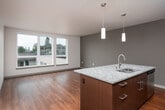 This single-floor residence showcases a contemporary open-concept layout that seamlessly connects the kitchen and living area, creating a spacious and functional living environment. Rich hardwood flooring flows throughout the space, lending warmth and continuity to every room.

The kitchen is a standout feature, outfitted with dark espresso cabinetry extending to ceiling height and complemented by a glass mosaic tile backsplash. Light granite countertops provide generous workspace, and a central island with matching granite offers additional prep space and casual seating. Stainless steel appliances—including a refrigerator, range, and over-the-range microwave—complete the polished, modern aesthetic. Pendant lighting and a track lighting system add focused illumination throughout.

The open living area is bathed in natural light courtesy of large picture windows that frame views of the surrounding neighborhood's greenery and urban landscape, contributing to a bright and airy atmosphere.

The bathroom is clean and well-appointed, featuring hardwood-style flooring, a granite-topped dark vanity, and a full tub-shower combination with white tile surround and chrome fixtures.

Overall, the space presents a cohesive modern design characterized by quality finishes, thoughtful lighting, and a layout that maximizes both openness and practicality.