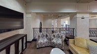 This lounge showcases a sophisticated multi-level layout connected by a broad, curved staircase with sleek dark handrails and clean-edged steps. The lower level features patterned carpet flooring in warm neutral tones, complemented by recessed lighting set within a distinctive dark-painted tray ceiling detail. Decorative bas-relief wall panels and curated canvas artwork add refined character throughout the space.

The upper level is defined by ornate wrought-iron railings with scroll detailing and warm wood-capped handrails, offering an open view to the levels below. Stone-tiled wall cladding in soft beige tones lends a classic, upscale ambiance. Arched doorway accents reinforce the architectural elegance.

Seating areas are thoughtfully arranged throughout, featuring patterned upholstered armchairs, a curved sofa, and occasional tables to encourage gathering. A wall-mounted flat-screen television with a dark wood media console provides an entertainment focal point in one seating area. A bar or service area is visible in the background, adding to the lounge's social functionality. Elevator access is also present, enhancing accessibility throughout the space.