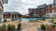 This expansive outdoor pool area is designed with a resort-style aesthetic, centered around a generously sized swimming pool with striking turquoise water. The pool features built-in tanning ledges, in-pool seating tables, and multiple water jet features that add visual interest and a dynamic ambiance to the space. Wide, flagstone-paved decking surrounds the pool, providing ample room for relaxation and circulation. Teal-cushioned lounge chairs are positioned throughout for sunbathing and leisure.

A brick pool house with floor-to-ceiling glass garage-style doors anchors one side of the pool area, offering both indoor and outdoor views of the water. Adjacent to the pool house, a wood pergola shelters an outdoor kitchen equipped with a built-in grill and stone countertops, making it well-suited for outdoor entertaining. A stone fire feature adds a focal point near the grilling area.

Lush tropical palms and carefully arranged landscaping with ornamental grasses and gravel beds are interspersed throughout, enhancing the resort atmosphere. The surrounding multi-story brick buildings frame the space, providing a sense of enclosure while the open sky above keeps the area feeling airy and expansive.