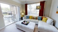 This single-floor residence offers a well-designed layout that balances functionality and comfort throughout its living spaces. The kitchen is thoughtfully arranged with white cabinetry extending to the upper walls, providing ample storage alongside a full suite of white appliances. A curved breakfast bar with light countertops serves as both a workspace and a casual dining spot, with bar stools positioned for easy seating. Recessed and overhead lighting keeps the kitchen bright and practical. An open pass-through connects the kitchen seamlessly to the rest of the living area, enhancing the sense of openness. The living room is a standout feature, filled with natural light streaming through large windows dressed in rich rust-colored drapes. Crown molding adds a refined architectural detail along the ceiling. A sliding glass door opens directly to a private balcony, offering outdoor access and additional natural light. The space is finished with soft carpeting underfoot, contributing to a warm and inviting ambiance. Together, the rooms create a cohesive, light-filled living environment that is both practical and comfortable.