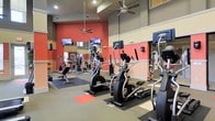 This fitness center is designed to provide a comprehensive workout experience within a single, open-plan floor. The space is defined by its bold, energizing color scheme, featuring vivid red-orange accent walls complemented by neutral tan and gray tones that create a motivating yet comfortable atmosphere. Ceiling fans are positioned throughout to ensure consistent airflow during workouts.

The cardio section is prominently featured along one side of the room, housing a row of elliptical trainers and stationary bikes, each equipped with individual monitor screens for entertainment or workout tracking. Multiple wall-mounted flat-screen TVs are installed at eye level, providing additional viewing options across the floor.

The strength training area occupies the rear of the space, outfitted with a multi-station cable and weight machine setup, adjustable benches, and free weight equipment. Protective rubber mats are laid beneath the equipment zones, clearly delineating workout areas from the main flooring. Large mirrors line portions of the walls, enhancing the sense of openness and allowing users to monitor their form. The overall layout is thoughtfully organized to allow for smooth traffic flow between cardio and strength training zones, making this fitness center a functional and well-appointed facility.