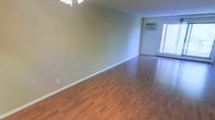 This single-floor space offers a clean, functional layout centered around an open living area. The room features warm-toned laminate hardwood flooring that runs throughout, providing a cohesive and low-maintenance surface underfoot. Neutral, light green-gray walls create a calm and versatile backdrop, well-suited for a variety of furnishing styles.

Climate control is well-addressed, with a wall-mounted air conditioning unit installed near the window and baseboard heating running along the perimeter — ensuring comfort across all seasons. A large window on the far wall allows natural light to filter into the space, brightening the interior and offering a connection to the outdoors.

The ceiling features a ceiling fan fixture, adding both airflow and a subtle design element overhead. White baseboards provide a clean contrast against the flooring and walls, giving the space a tidy, finished appearance. The open, unobstructed layout offers flexibility in how the space can be arranged and utilized, making it a practical and adaptable living environment.