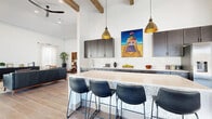 This well-appointed single-story space combines contemporary design with warm, inviting details throughout its open layout. The kitchen serves as a central focal point, showcasing dark charcoal cabinetry, a patterned tile backsplash, and a generous island with a light quartz countertop accented by decorative mosaic tile siding. Leather-upholstered counter stools line the island, while a pair of hammered brass pendant lights hang overhead. A stainless steel refrigerator and ample upper and lower cabinetry complete the functional cooking space. Vibrant artwork adds a bold splash of color against the white walls.

The lounge areas benefit from soaring ceilings with exposed wooden beams and wood-look tile flooring throughout. One seating zone features a round dining table alongside swivel chairs, bathed in natural light from large windows dressed with sheer curtains. A separate lounge opens directly to an outdoor terrace through full-height sliding glass doors, offering sweeping views of the surrounding community and pool area.

A dedicated office nook provides leather seating and a sleek credenza beside floor-to-ceiling glass doors. The accessible restroom is finished with large-format wall tile, wood-look flooring, a floating vanity, a round gold-framed mirror, and compliant grab bars throughout.