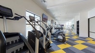 This fitness center is a generously proportioned, single-floor facility designed to accommodate a full range of workout needs. The space is organized into two primary zones: a dedicated cardio area and a strength training section, each clearly defined yet flowing seamlessly into one another.

The cardio zone runs along one side of the facility and features a substantial row of Life Fitness equipment, including treadmills, elliptical trainers, and stair climbers. Multiple wall-mounted flat-screen TVs are positioned throughout this zone, providing entertainment during workouts. A series of large windows lines the wall, flooding the area with natural light and offering views of the surrounding neighborhood.

The strength training area is equally well-appointed, housing a selection of cable machines, multi-use weight stations, a dumbbell rack, and adjustable benches. Floor-to-ceiling mirrors along one wall visually expand the space and allow users to monitor their form. Tall windows draw in additional natural light in this section.

Throughout the facility, a bold checkered carpet in yellow, grey, and charcoal adds a distinctive, energetic aesthetic. The white walls and clean architectural lines contribute to a bright, modern ambiance, making this fitness center both functional and visually engaging.