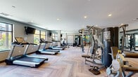 This fitness center is organized into distinct training zones, offering a comprehensive workout environment suitable for a wide range of exercise routines. The main cardio area features a row of treadmills and elliptical machines positioned along large windows that invite natural light and provide views of the surrounding outdoor greenery. Wall-mounted televisions are stationed throughout this section, enhancing the cardio experience. The flooring in this zone showcases a herringbone-patterned wood-look surface that adds a contemporary aesthetic to the space.

Adjacent to the cardio area, a strength training zone houses a Jones machine, a cable pull system, free weight benches, and additional resistance equipment, all reflected in floor-to-ceiling mirrors that amplify the sense of space. Recessed lighting throughout maintains a bright, consistent atmosphere.

A separate functional training room features cork-style flooring, large glass doors opening to the outdoors, and a full wall of mirrors. This space is equipped with spin bikes, stability balls, resistance bands, kettlebells, step platforms, and a large flat-screen display, making it well-suited for group fitness or individual cross-training sessions.