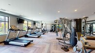 This fitness center is organized into distinct training zones, offering a comprehensive workout environment suitable for a wide range of exercise routines. The main cardio area features a row of treadmills and elliptical machines positioned along large windows that invite natural light and provide views of the surrounding outdoor greenery. Wall-mounted televisions are stationed throughout this section, enhancing the cardio experience. The flooring in this zone showcases a herringbone-patterned wood-look surface that adds a contemporary aesthetic to the space.

Adjacent to the cardio area, a strength training zone houses a Jones machine, a cable pull system, free weight benches, and additional resistance equipment, all reflected in floor-to-ceiling mirrors that amplify the sense of space. Recessed lighting throughout maintains a bright, consistent atmosphere.

A separate functional training room features cork-style flooring, large glass doors opening to the outdoors, and a full wall of mirrors. This space is equipped with spin bikes, stability balls, resistance bands, kettlebells, step platforms, and a large flat-screen display, making it well-suited for group fitness or individual cross-training sessions.