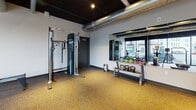 This modern fitness center is thoughtfully designed to support a wide range of workout routines. The space features an industrial-style aesthetic, with exposed ductwork and dark-painted ceilings that contrast sharply against crisp white walls, giving the gym a contemporary, urban character. Durable rubber flooring in a speckled gold tone covers the entire floor, providing both cushioning and traction during exercise.

Large floor-to-ceiling windows line one side of the space, offering expansive street-level views and allowing abundant natural light to permeate the interior, creating an open and energizing atmosphere. The cardio zone is well-appointed with multiple treadmills, elliptical trainers, and a stationary bike, all positioned to take advantage of the street views.

The strength and functional training area is equally well-stocked, featuring a multi-cable functional trainer, a full rack of dumbbells in varying weights, kettlebells of multiple sizes arranged on a dedicated rack, medicine balls, foam rollers, and a balance board. A weight bench provides additional versatility for strength exercises. Recessed spotlights and track lighting ensure the entire space is well-illuminated. The gym is accessed through a wood-paneled door and includes clearly marked emergency exit signage, reflecting attention to safety and functionality throughout.