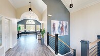 Located at 7001 W Cross Creek Trail in Brecksville, Ohio, this two-floor community space offers a generous and well-appointed layout across two distinct levels. The main floor greets visitors with a striking hallway and staircase area defined by soaring vaulted ceilings, bold slate-blue accent walls, rich wood-toned luxury vinyl plank flooring, and a sleek modern staircase featuring black metal railings with warm wood handrails. Large-scale artwork and pendant lighting add a sophisticated touch to the space. The adjacent living and dining area is bathed in natural light from expansive windows, including a dramatic arched window that frames outdoor views of lush, colorful foliage. Vaulted ceilings and deep blue walls lend this room an airy, refined ambiance with bistro-style seating for casual gatherings. A separate living room offers a cozy fireplace, crown molding, patterned carpet, and ample seating for relaxed conversation. The kitchen is outfitted with gray shaker-style cabinetry, white countertops, stainless steel appliances, and black hardware accents. The lower level houses a well-equipped exercise room featuring treadmills, ellipticals, free weights, an adjustable bench, floor-to-ceiling mirrors, rubber flooring, and access to restroom facilities — providing a comprehensive fitness experience within the property.
